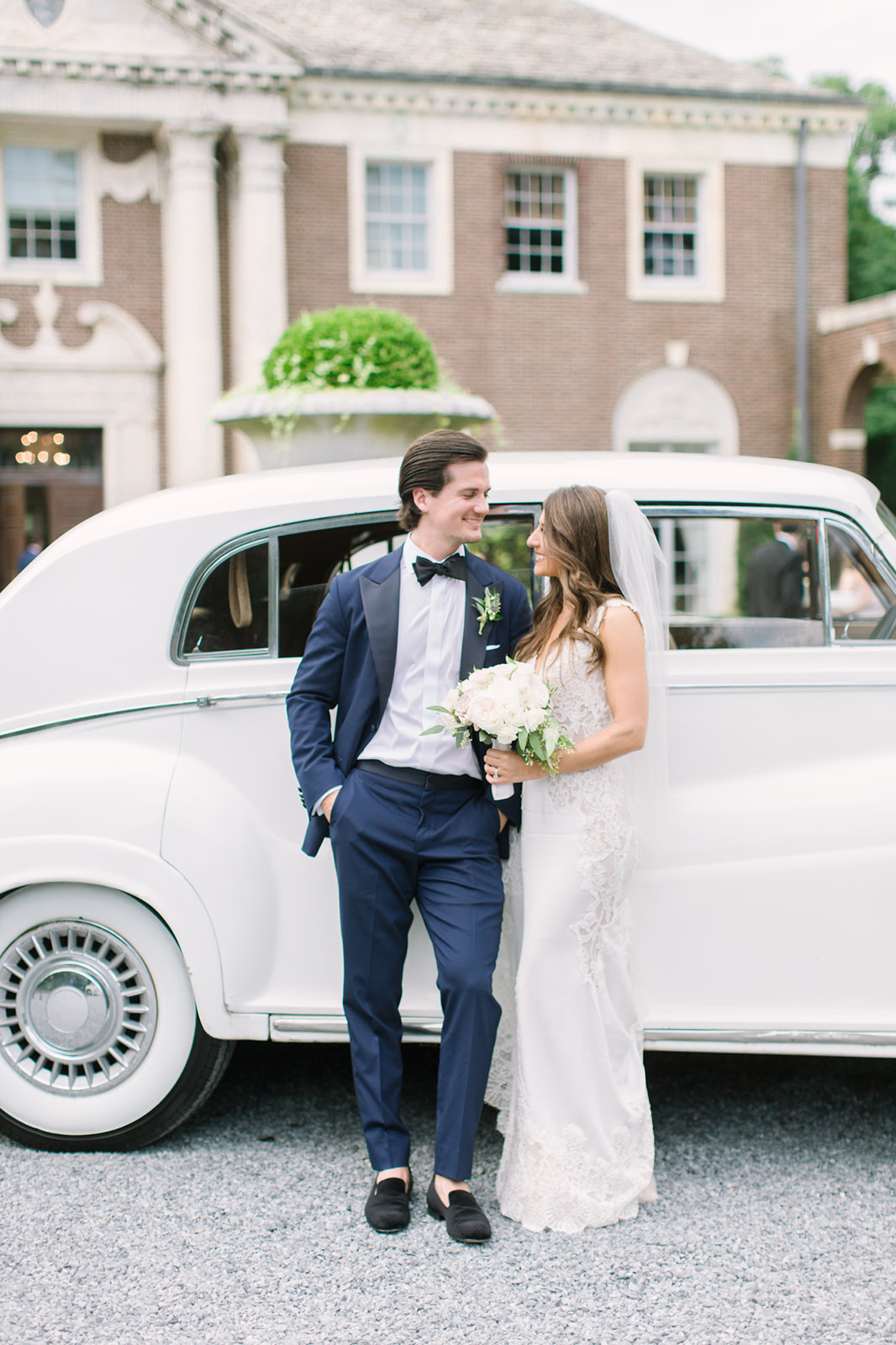 Bride and Groom posing next to a white Rolls Royce car. They are both smiling at each other .